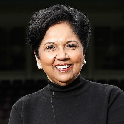 A person with short black hair, wearing a black turtleneck and pearl earrings, is smiling warmly, exuding the charm of a curated customer experience. The background is dark and out of focus.