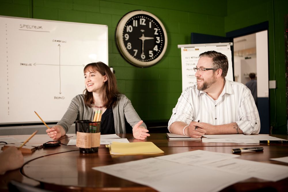 Two people are sitting at a round table in a meeting room with a green wall. A clock and whiteboards with charts illustrating customer experience improvements are visible in the background. They are smiling, with papers and pens on the table, discussing innovative digital solutions.