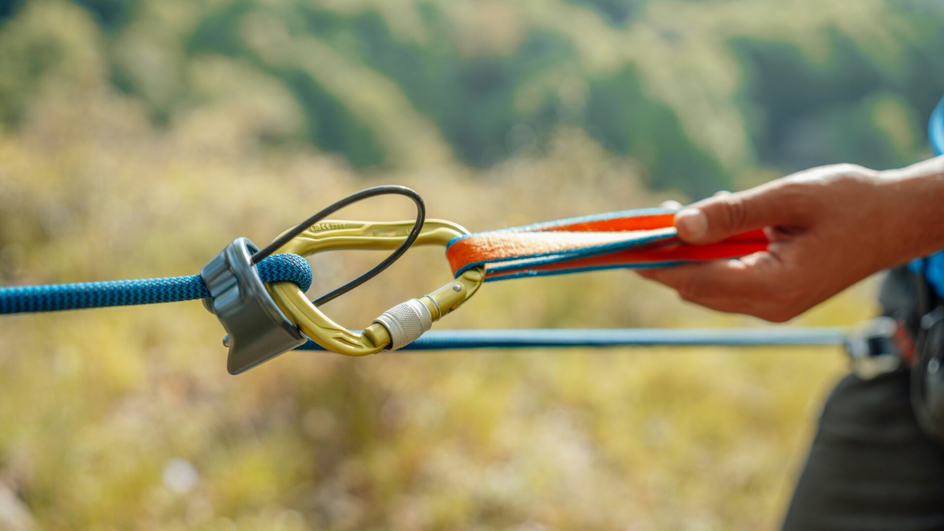 A close-up of a hand holding climbing gear, including a carabiner attached to blue and orange ropes—acting as the essential middleware connecting each part—set against a blurred outdoor background.