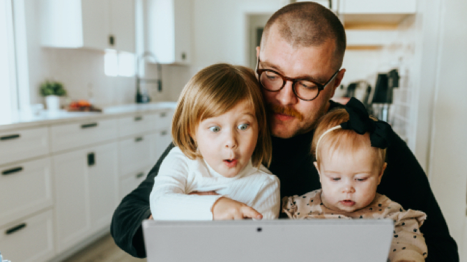 A father with glasses sits in a kitchen holding two young children on his lap. The older child looks surprised while looking at a laptop screen, intrigued by the delightful user experience, while the younger child, with a bow in their hair, gazes down at the device.