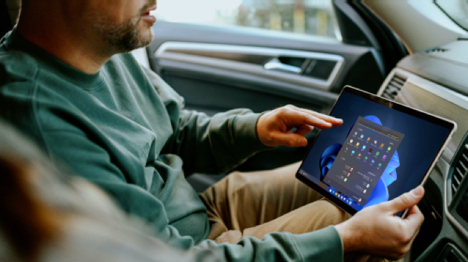 A person in a green sweater sits in a car, using a tablet displaying a grid of colorful app icons. The user experience is enhanced by the dark-themed menu as they touch the screen with their right hand, seamlessly navigating through options.