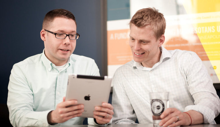 Two men are sitting and discussing user experience while looking at an iPad together. One holds the tablet, and the other has a glass of water. Both wear light-colored shirts, with a blurred poster in the background serving as a subtle nod to refined design principles.