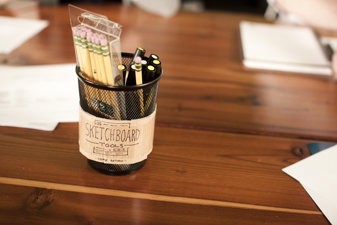 On a wooden table, the mesh holder labeled Sketchboard Tools is brimming with pencils and markers wrapped in paper. Papers are scattered about like Clockwork creations, each vying to enhance the customer experience.