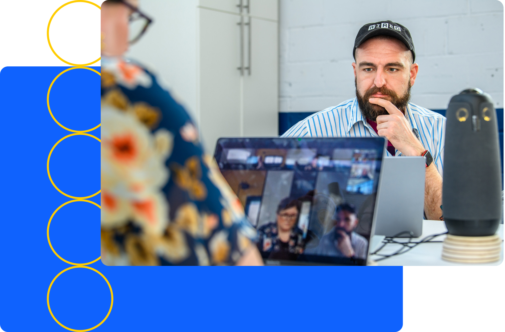 A man in a cap sits at a table with a laptop, engaging in a video call about user experience design. Another person in a floral shirt is partially visible. A device stands nearby against the white brick wall with blue accents, creating an inviting setting for creative collaboration.