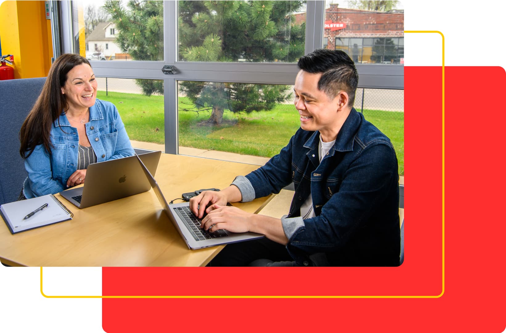 Two people are sitting at a table, each with a laptop, smiling and engaged in conversation about custom software. They are in a bright room with large windows showing a green outdoor area. A red and yellow graphic border frames part of the image.