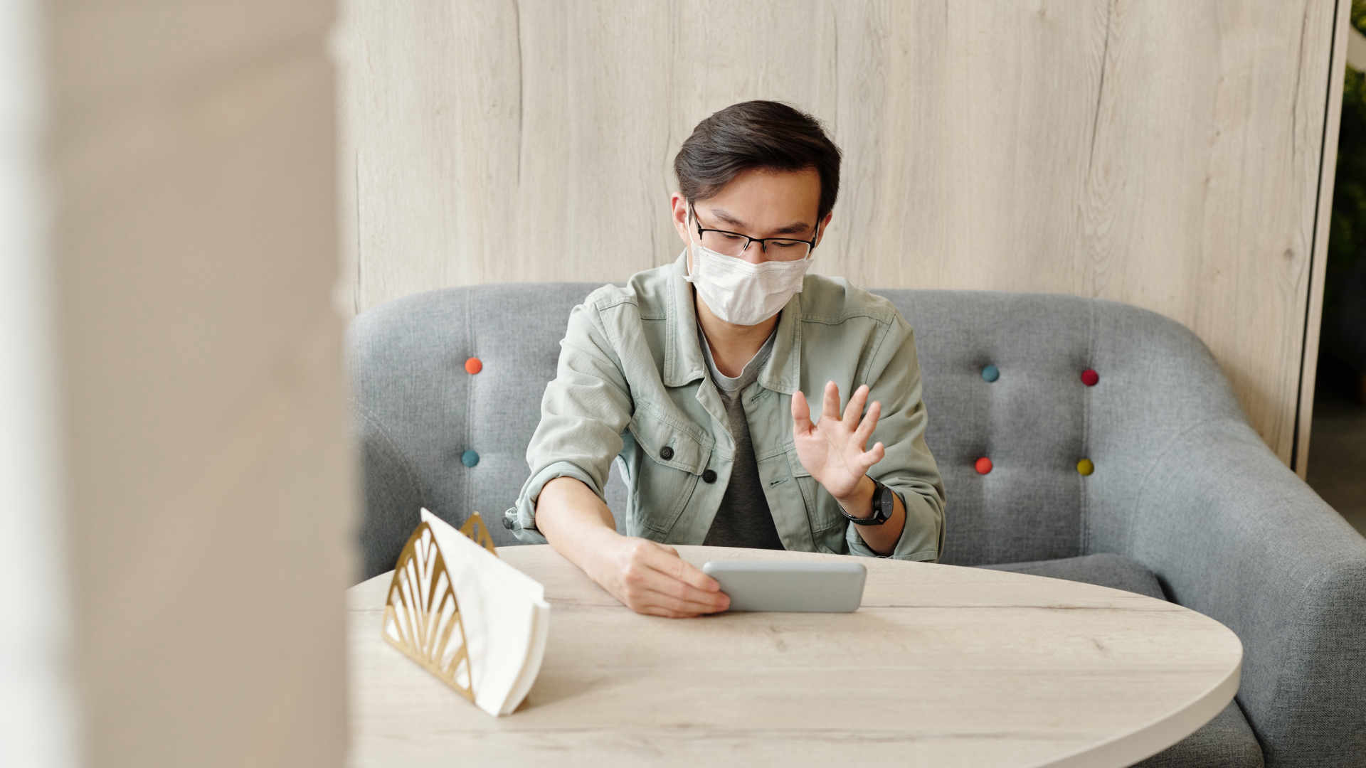 A person wearing glasses and a face mask is sitting on a gray sofa, waving at a tablet on a round table—engaging with a digital solution. The background features a light wooden wall and a colorful pillow, enhancing the user experience.