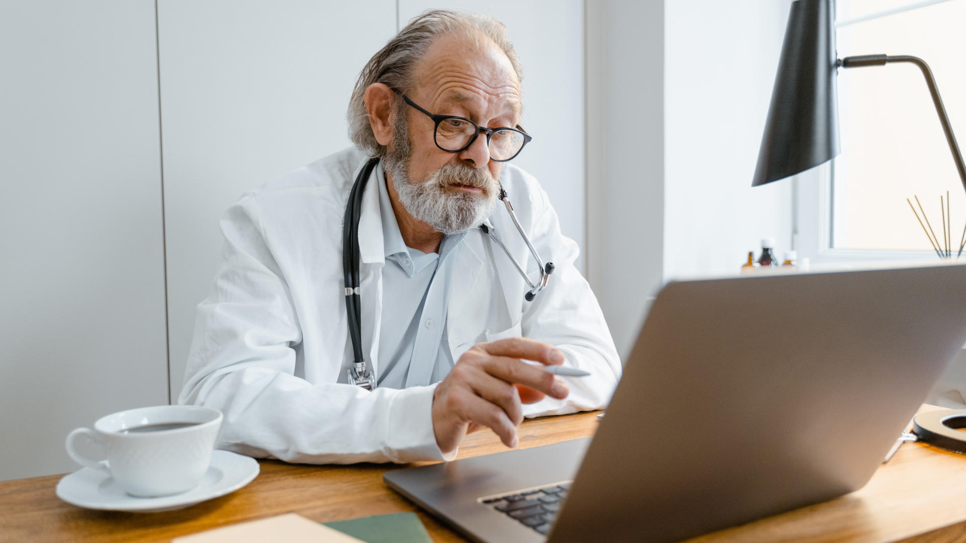 Elderly man with glasses and a stethoscope around his neck, wearing a white coat, sits at a desk working on a laptop using custom software. A cup of coffee rests beside him, and a modern lamp glows in the background.
