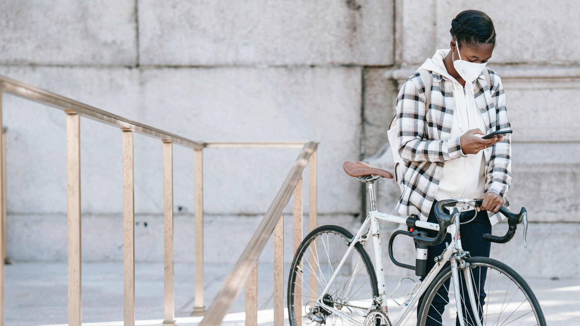 A person in a plaid jacket and face mask stands beside a white bicycle, engaged with their smartphone like clockwork. They are next to a metal railing on a stone-paved surface, with a large stone wall standing impressively in the background.