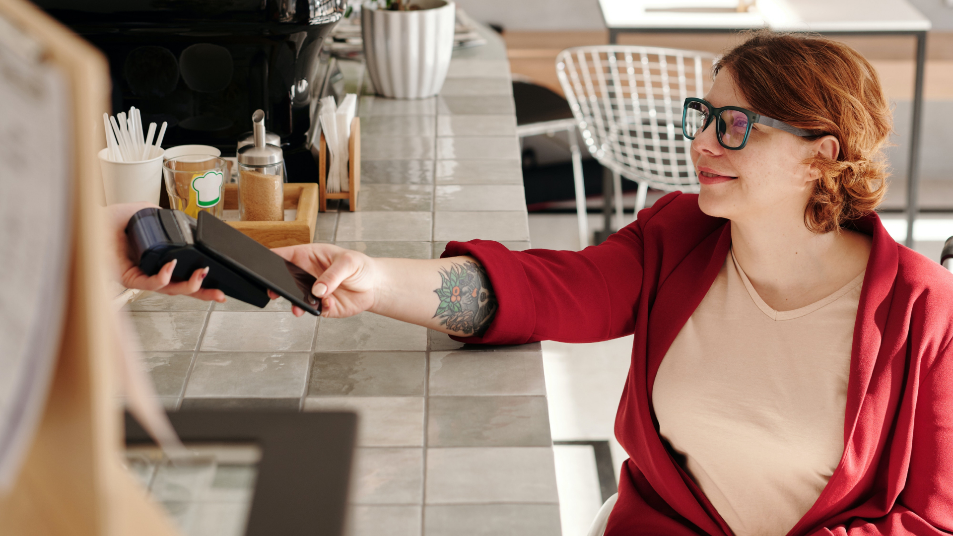 A person with glasses and a red blazer is seated at a table, smiling while handing a card to a cashier holding a sleek payment terminal. The transaction proceeds with clockwork precision in the modern café, enhanced by custom software for an impeccable user experience.