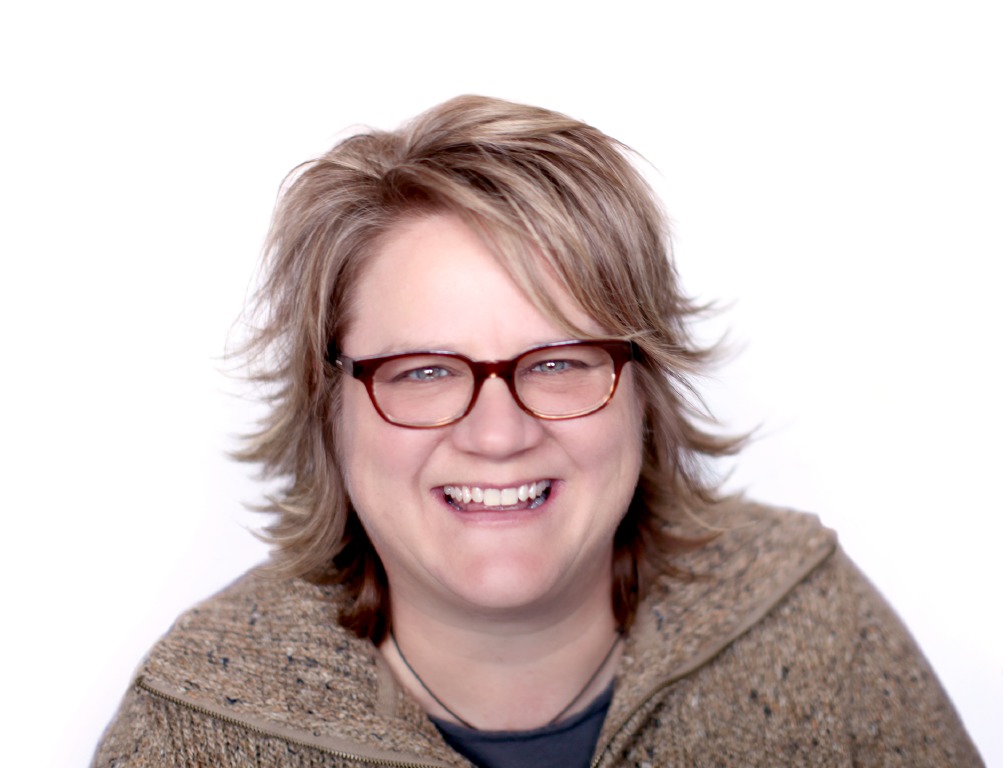 A person with short, tousled hair and glasses smiles at the camera, exuding a clockwork charm. They wear a speckled sweater with a zipper against a plain white background, creating an effortless blend of warmth and style.