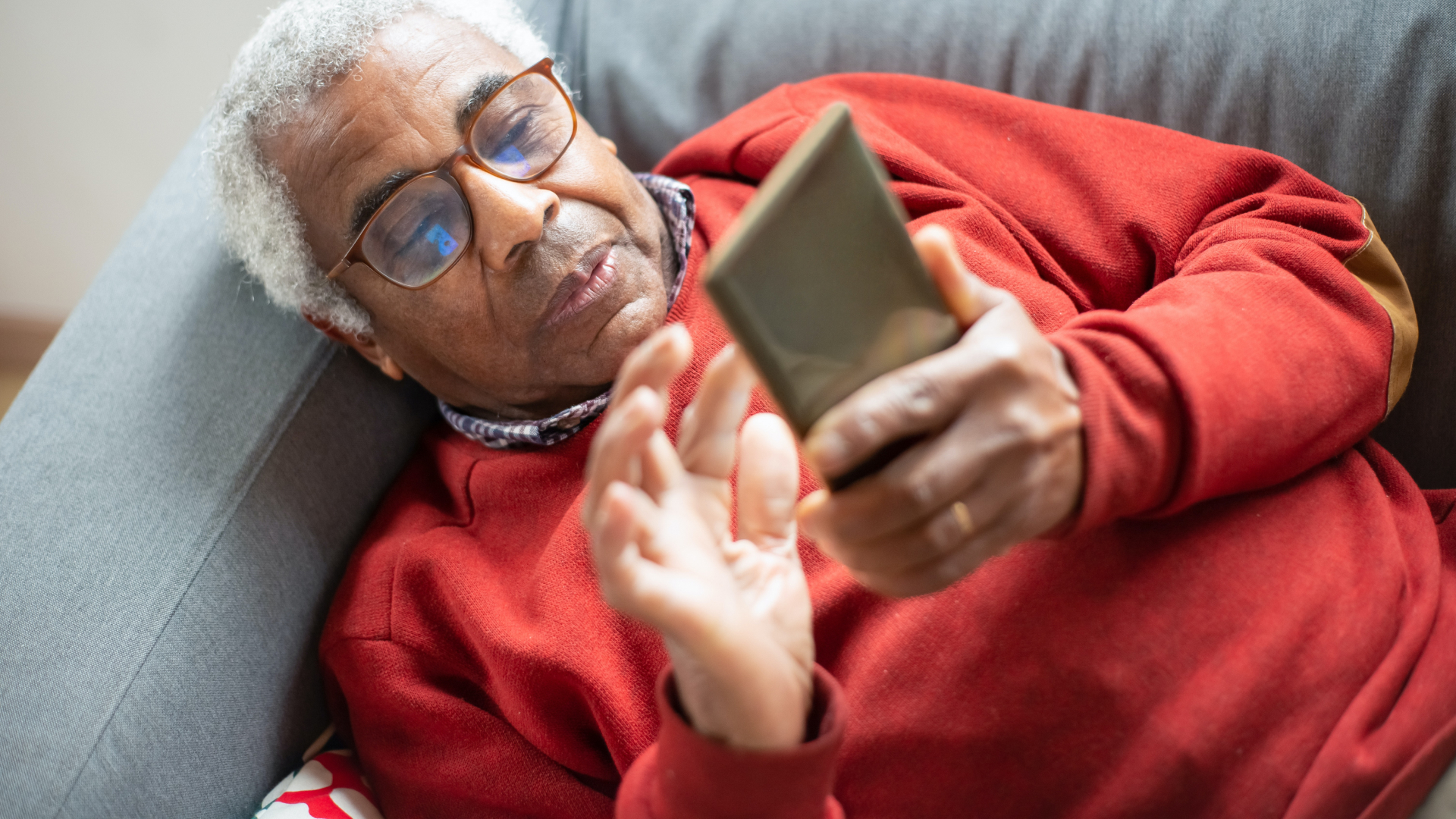 An elderly man with gray hair and glasses, donning a red sweater, is comfortably lying on a gray couch, enhancing his user experience by navigating through his smartphone with ease.