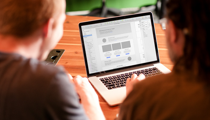 Two people sit at a wooden table, immersed in experience design, as they focus on a laptop displaying a website mockup. The screen reveals a layout with text and image placeholders. The background is blurred, emphasizing their clockwork collaboration on enhancing customer experience.