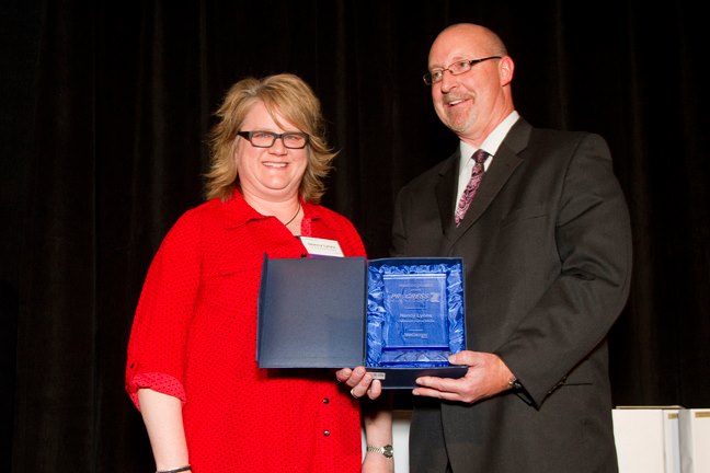 A woman in a red blouse and a man in a suit smile while holding a blue award plaque together on stage, celebrating their innovative digital solution. The dark background adds to the sense of achievement.