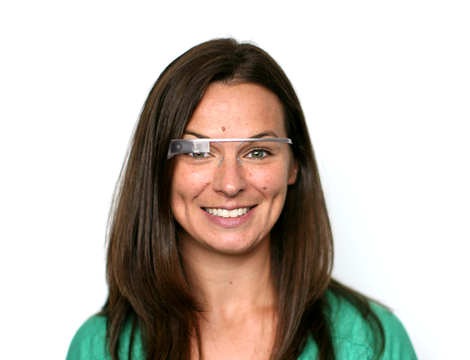 A woman with long brown hair is smiling while wearing smart glasses, embodying a seamless digital solution. She stands against a plain white background, her green top adding a touch of vibrancy to the scene.
