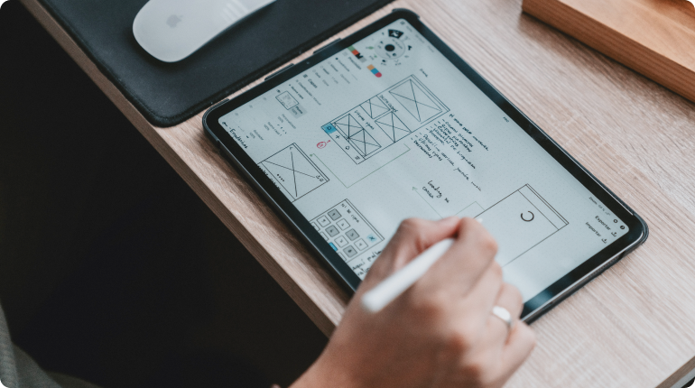 A person uses a stylus on a tablet showcasing wireframe designs and notes, reflecting a seamless user experience. Nearby, a computer mouse rests on its pad, all set upon the wooden desk as part of an efficient digital solution.
