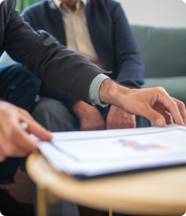 A person in a suit is pointing at a document on a table, highlighting a proposed custom software solution, while another individual sits in the background on a couch. The focus is on the hand and the document, underscoring an engaging discussion about enhancing customer experience.