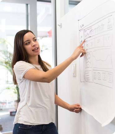 A woman in a white shirt presents a large sheet of paper with charts and diagrams on a whiteboard, pointing to emphasize insights on customer experience. The blurred office environment in the background suggests a focus on enhancing strategic decisions.