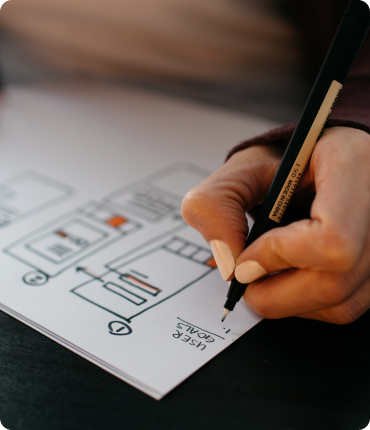 Close-up of a person sketching a design layout on paper with a marker. The paper includes flowchart-like diagrams and the words USER GOALS, capturing the essence of experience design. The persons hand is visible, focused on crafting an innovative digital solution.