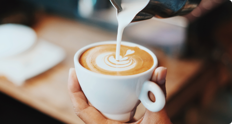 A close-up of a hand holding a white cup as milk is poured to craft latte art, akin to experience design on coffee. The background is softly blurred, drawing attention to the swirling patterns adorning the top.
