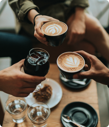 Three people clink coffee cups with latte art over a table adorned with glasses of water and a pastry, reminiscent of the seamless precision of Clockwork. One person holds a glass of iced coffee, creating a scene that is both relaxed and social, enhancing the overall user experience.
