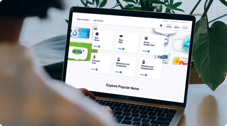A person is using a laptop to explore an online healthcare store, seamlessly navigating through an expertly crafted user experience. Categories like Stay Well and Personal Care are displayed on the screen alongside products such as sanitizers and masks, with lush plants in the background enhancing the digital solution.