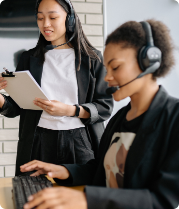 Two women wearing headsets are working. One is seated, typing on a keyboard, while the other, standing with a clipboard and pen, discusses the latest digital solution. Both dressed in black blazers, they appear to be engaged in enhancing the customer experience.