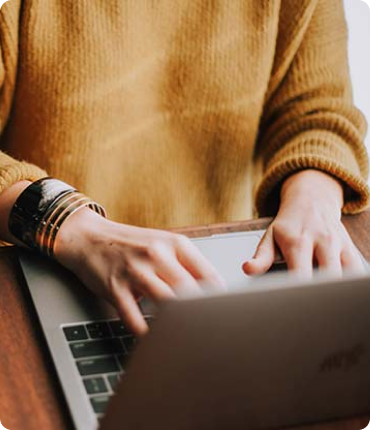 A person in a mustard sweater is typing on a laptop at a wooden desk, wearing multiple bracelets on the left wrist, as they focus intently on crafting seamless user experiences like clockwork.
