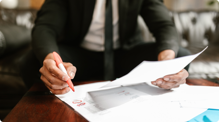 A person in a suit is sitting on a sofa, meticulously marking a document with a pen. The focus is on the hand expertly handling the paperwork as if orchestrating intricate Clockwork, ensuring every detail aligns in this digital solution of precision and intent.