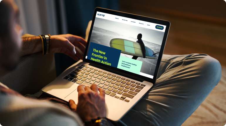 A person sits with a laptop on their lap, exploring a website titled The New Frontier in Health Action. Like clockwork, the digital solution showcases a surfer holding a surfboard on a beach. The room is dimly lit, and the person wears a brown sweater.