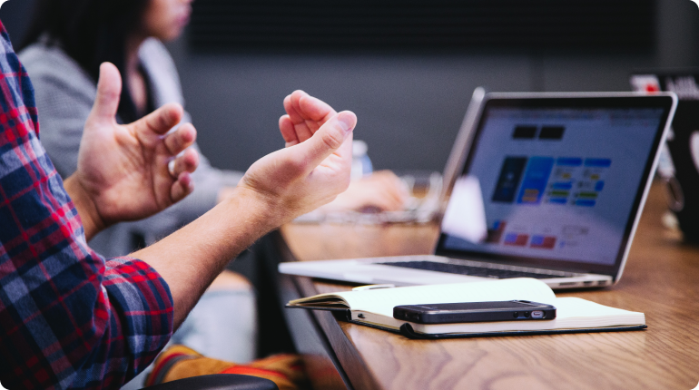 A person in a plaid shirt gestures animatedly, emphasizing the importance of customer experience during a discussion. An open laptop displaying a colorful chart sits on the wooden table, accompanied by a notebook and smartphone. Another person is partially visible in the background.