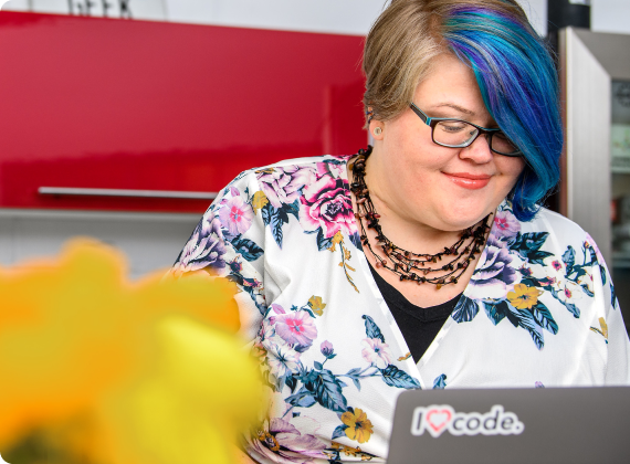 A person with short blue and brown hair, wearing glasses and a floral top, smiles while looking at a laptop adorned with an I ♥︎ code sticker. Red cabinets are visible in the background, as yellow flowers blur in the foreground—a scene reflecting seamless user experience akin to clockwork.