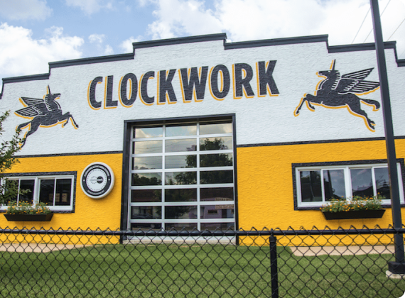 A building with a white and yellow façade features a large CLOCKWORK sign and images of winged horses. Enhancing user experience, there are windows with flower boxes, a glass garage door, and a grassy area behind a black fence in the foreground.
