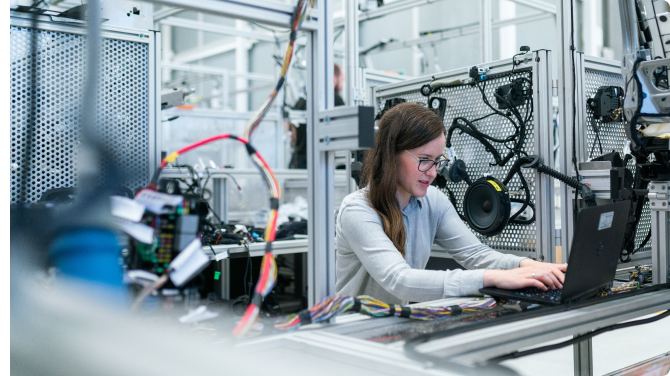 A woman with long hair is working on a laptop in a high-tech lab, focused on creating an innovative digital solution. Surrounded by electronic equipment, wires, and metal structures, she embodies the cutting-edge world of programming and engineering.