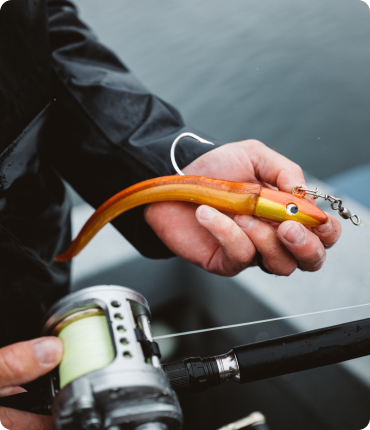 A person in a dark rain jacket holds a fishing rod with a reel in one hand and a bright orange eel-shaped fishing lure with a metal hook in the other, capturing the essence of seamless user experience. The background shows blurred water.