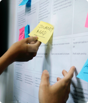 Two hands place sticky notes on a board filled with paper sections. One yellow note reads Journey Map, pointing to an experience design session. Blue and pink notes are partially visible, suggesting a planning or brainstorming session focused on enhancing user experience.