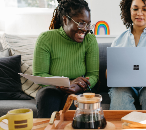 Two people sit on a couch, smiling at a laptop like they’ve nailed the art of customer experience. One, with long hair and papers in hand, dons a green top. The other sports a blue shirt and curly hair. Nearby sits a glass coffee pot and rainbow decoration, adding to the serene atmosphere.