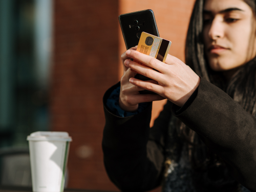 A person holds a smartphone and credit card outdoors, focused on enhancing their user experience through an online transaction. A takeaway coffee cup rests on the table beside them, with a brick wall and blurred background adding context to this seamless customer experience.