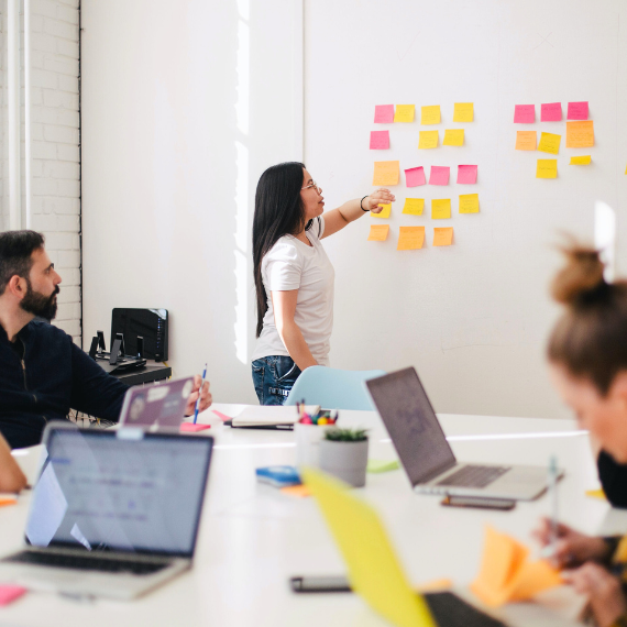 A woman stands by a whiteboard covered in colorful sticky notes, explaining an experience design strategy. Two people sit at a table with open laptops and documents. The room is well-lit with natural light from a window, resembling the meticulous precision of Clockwork.