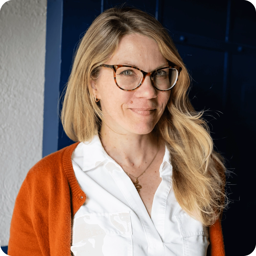 Jaden Johnston, Clockwork. A woman with long blonde hair and glasses, dressed in a white shirt and orange cardigan, stands in front of a navy blue door, smiling softly at the camera—reflecting the approachable professionalism of technology consultants.
