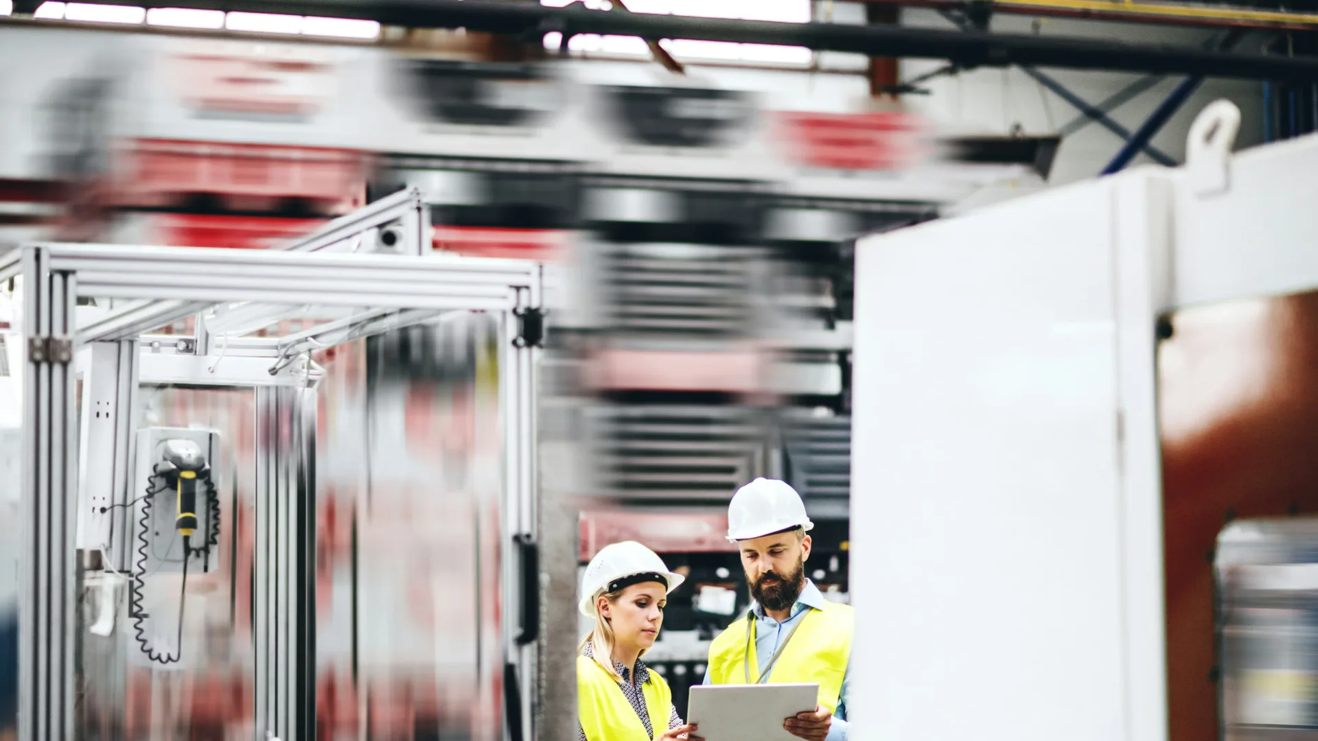 Two workers in safety vests and helmets stand in a busy factory, looking at a tablet and discussing something, with blurred machinery and shelves in the background.